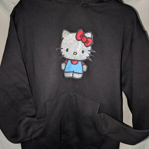 May include: Black hoodie with a Hello Kitty design. The graphic shows Hello Kitty with a red bow, blue overalls, and a rhinestone-like design. The sweatshirt has a front pocket and a drawstring hood.