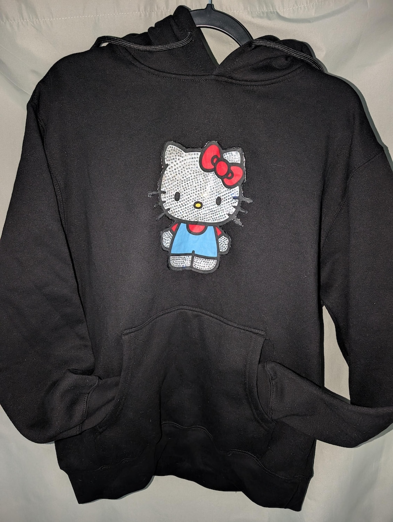 May include: Black hoodie with a Hello Kitty design. The graphic shows Hello Kitty with a red bow, blue overalls, and a rhinestone-like design. The sweatshirt has a front pocket and a drawstring hood.