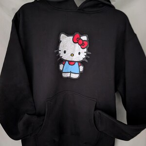 May include: Black hooded sweatshirt with a Hello Kitty patch. The patch depicts Hello Kitty with a red bow, blue overalls, and a white body. The sweatshirt has a front pocket and a drawstring hood.