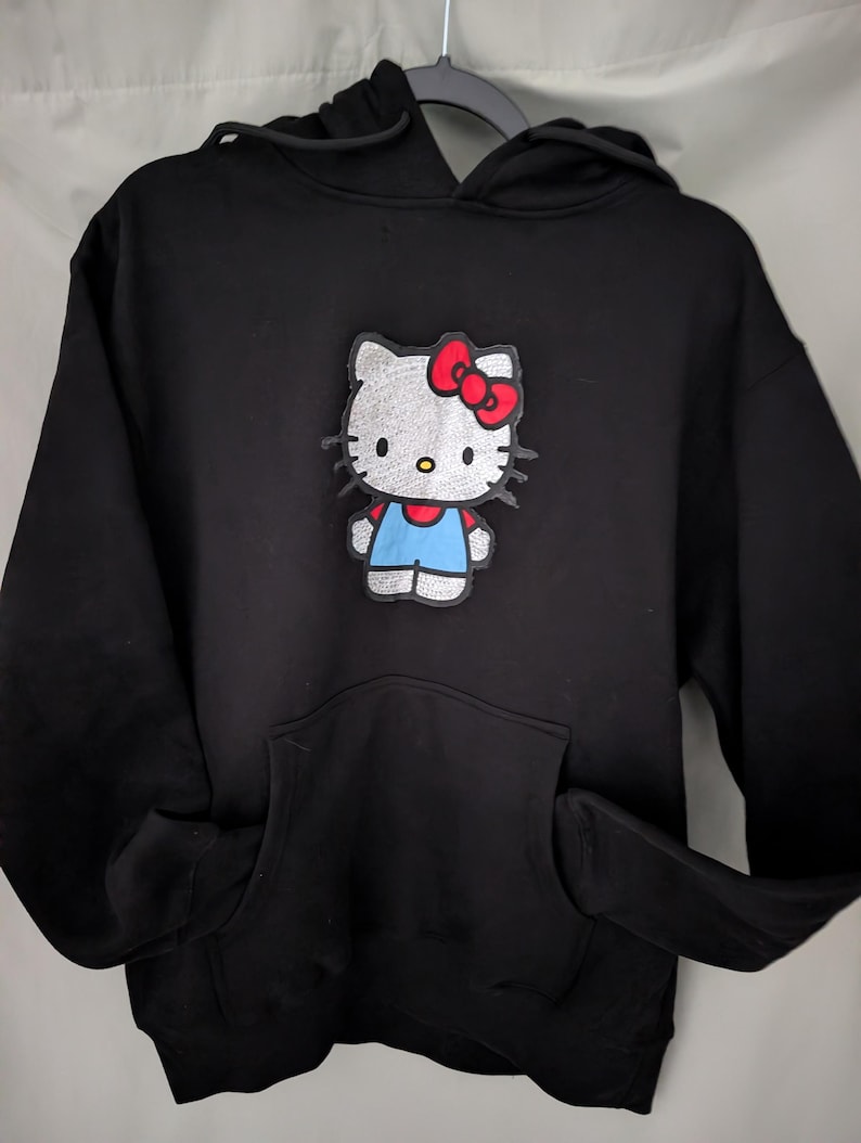 May include: Black hooded sweatshirt with a Hello Kitty patch. The patch depicts Hello Kitty with a red bow, blue overalls, and a white body. The sweatshirt has a front pocket and a drawstring hood.