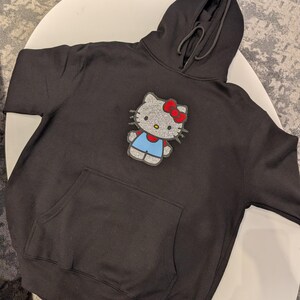 May include: Black hooded sweatshirt featuring a large, sparkling Hello Kitty design. The design includes a red bow, blue overalls, and a silver face. The sweatshirt has a front pocket and a drawstring hood.