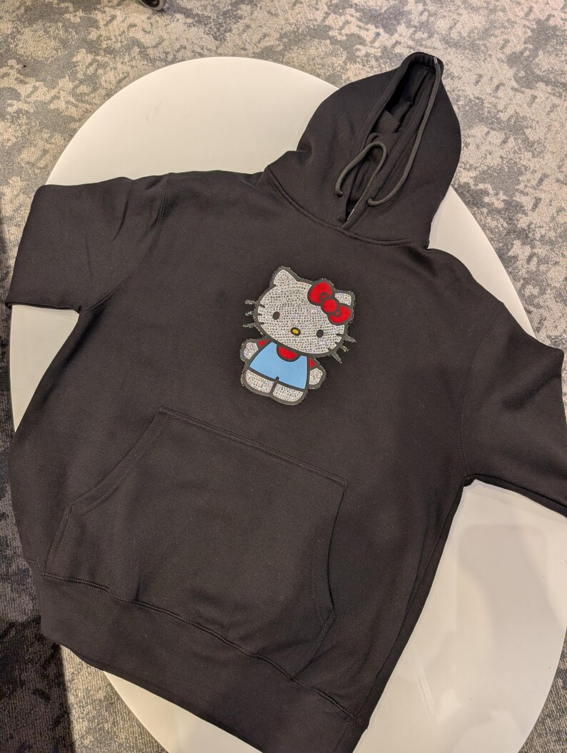 May include: Black hooded sweatshirt featuring a large, sparkling Hello Kitty design. The design includes a red bow, blue overalls, and a silver face. The sweatshirt has a front pocket and a drawstring hood.