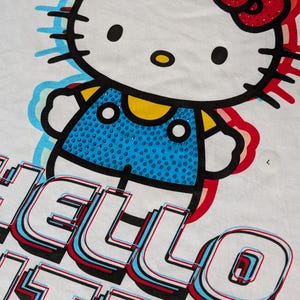 May include: White t-shirt with a colorful graphic of Hello Kitty wearing a blue overall with rhinestones. The text "Hello Kitty" is printed in a 3D style with red, blue, and white accents.