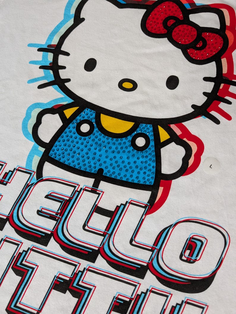 May include: White t-shirt with a colorful graphic of Hello Kitty wearing a blue overall with rhinestones. The text "Hello Kitty" is printed in a 3D style with red, blue, and white accents.