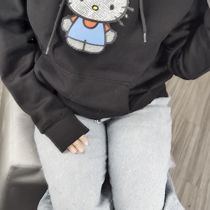May include: Black hooded sweatshirt with a Hello Kitty design, featuring a red bow and blue overalls. The sweatshirt has a front pocket and a drawstring hood. The person is wearing light grey jeans.