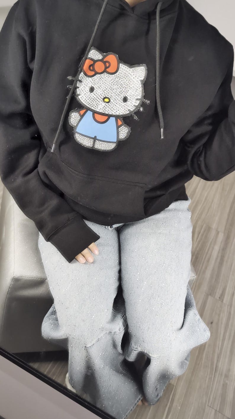 May include: Black hooded sweatshirt with a Hello Kitty design, featuring a red bow and blue overalls. The sweatshirt has a front pocket and a drawstring hood. The person is wearing light grey jeans.