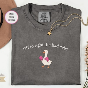 May include: A charcoal gray t-shirt featuring the white text "Off to fight the bad cells" above a graphic of a white goose wearing pink boxing gloves and a small pink ribbon on its chest. The shirt has a Comfort Colors brand label at the neck.