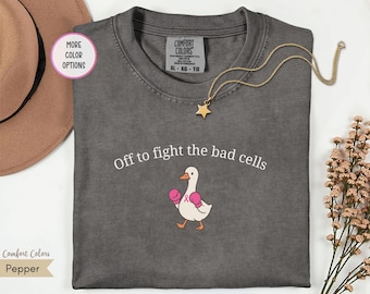 Puede incluir: Una camiseta gris carbón con el texto blanco "Off to fight the bad cells" sobre un gráfico de un ganso blanco con guantes de boxeo rosas y un pequeño lazo rosa en el pecho. La camiseta tiene una etiqueta de la marca Comfort Colors.