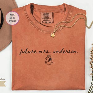 May include: A yam-colored crew neck t-shirt featuring the black text future mrs. anderson and a small line drawing of a ghost holding a bouquet of flowers. The shirt has a Comfort Colors brand label on the inside collar indicating size XL.