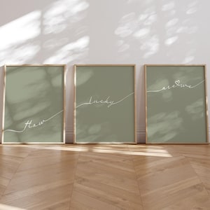 Op de afbeelding: Drie ingelijste prints met een saliegroene achtergrond en witte cursieve tekst. De tekst op de prints luidt "How", "Lucky" en "Are We" met een hartje.