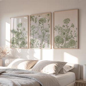 Può includere: Tre stampe botaniche incorniciate con illustrazioni floreali verdi. Le stampe sono esposte sopra un letto con biancheria da letto dai colori neutri. Le cornici sono di colore legno chiaro e l'opera presenta disegni dettagliati di vari fiori e fogliame.