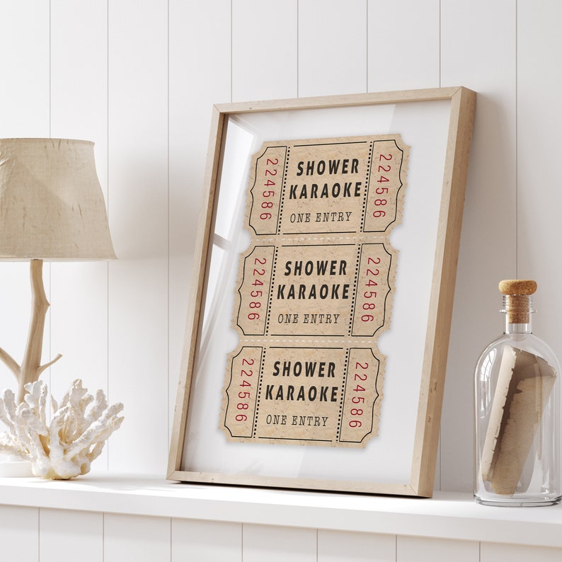 May include: Three vintage-style tickets with the text "Shower Karaoke" and "One Entry" printed on them. The tickets are framed in a light wood frame and displayed on a white shelf.