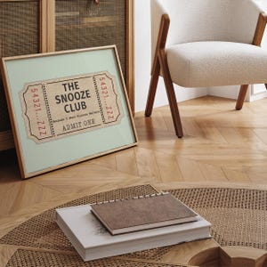 May include: A framed print of a vintage ticket with the text "The Snooze Club" and the words "Because 5 More Minutes Matters" and "Admit One". The ticket is printed on a light brown background with red and black text.
