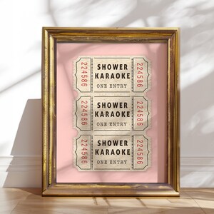 May include: Three vintage-style tickets with the text "Shower Karaoke" and the number "224586" printed on each. The tickets are displayed in a gold frame against a light pink background.