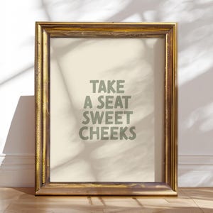 Könnte beinhalten: Gerahmter Druck mit dem Text "TAKE A SEAT SWEET CHEEKS" in grünen Buchstaben. Der Rahmen ist goldfarben mit einem Used-Look. Der Hintergrund ist hellbeige.