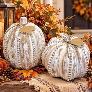 May include: Two decorative pumpkins featuring a distressed white and natural wood finish with beaded ridges. Each pumpkin is accented with a twine-tied tan paper leaf at the stem, showcasing a textured, rustic appearance with vertical ribbing.