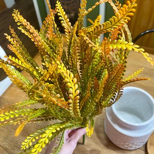 May include: A bundle of artificial fern stems in shades of yellow, green, and orange. The fern has a delicate, textured appearance. A white ceramic jar is visible in the background.