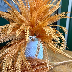 May include: A cluster of artificial fern-like stems in a metallic copper-orange hue, featuring numerous small, rounded leaves on thin branches. The stems are arranged in a white ceramic vase with a textured, diamond-patterned base.