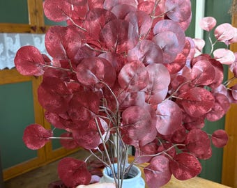 May include: A bundle of artificial eucalyptus stems with burgundy-colored, round leaves. The stems are held in a white ceramic vase. The leaves have a gradient effect, transitioning from a darker burgundy to a lighter pinkish hue.