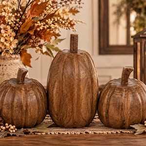 May include: Three brown pumpkin-shaped decorative objects with a textured, wood-like finish and prominent vertical ridges. The central pumpkin is taller and more elongated, while the two flanking pumpkins are shorter and rounder in shape.