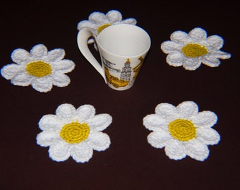 May include: A white ceramic mug with a yellow and black design sits among five white daisy-shaped crochet coasters with yellow centers. The coasters are arranged on a dark brown surface.
