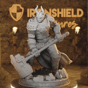 Puede incluir: Figura gris detallada de un guerrero con armadura completa, empuñando un gran martillo. La figura tiene un casco con cuernos y está posada sobre una base rocosa. El fondo presenta el texto "IRONSHIELD" y un logotipo de escudo.
