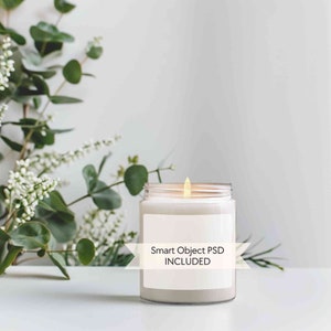 May include: A white candle in a clear glass jar with a white label that says "Smart Object PSD INCLUDED". The candle is lit and the flame is visible. The candle is on a white surface with green foliage in the background.