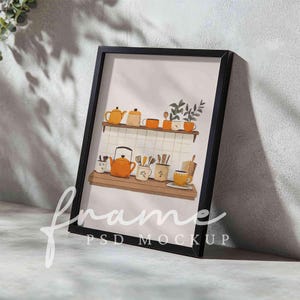 May include: A black framed print with a watercolor illustration of a kitchen shelf with orange teapots, mugs, and utensils. The print is on a white background and has a light brown wood shelf.