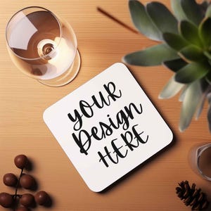 Op de afbeelding: Witte vierkante onderzetter met zwarte tekst die "your design here" zegt op een houten oppervlak.