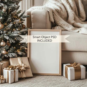 Peut inclure: Une maquette d'affiche encadrée blanche avec le texte "Smart Object PSD INCLUDED" sur un fond blanc. L'affiche est posée sur un tapis beige devant un canapé blanc avec une couverture en tricot beige. Il y a des cadeaux emballés avec un ruban doré et des nœuds devant l'affiche.