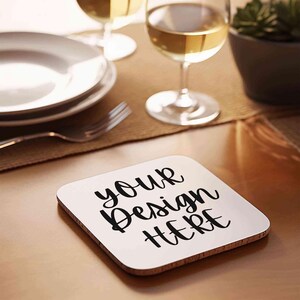 May include: White square coaster with the text "your design here" in black cursive font. The coaster is on a wooden table with a glass of white wine and a plate in the background.