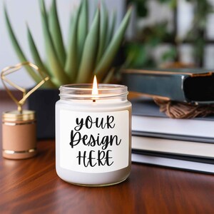 May include: A white candle in a clear glass jar with a white label that says "your design here". The candle is lit and the flame is visible. The candle is sitting on a wooden table with books and a plant in the background.