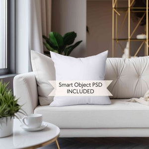 May include: White throw pillow mockup on a white couch with a white coffee table and a green plant. The pillow has a white background with the text "Smart Object PSD INCLUDED" in black.