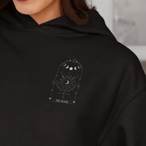 May include: Black hoodie with a front pocket and a hood. The design on the upper back features a celestial graphic with moons, stars, and a sun, along with the text "THE MOON".
