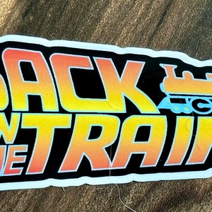 May include: A sticker with the words "BACK ON THE TRAIN" in a gradient yellow to orange font, outlined in black. A cartoon train in orange and blue is on the right. The sticker has a white border.