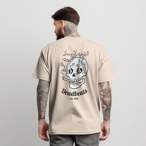 May include: A beige t-shirt with a black and white graphic of a skull smoking a cigarette. The text "Deadbeats Est. 2016" is printed below the skull.