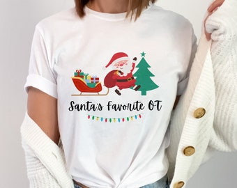 May include: White t-shirt with a cartoon image of Santa Claus in a red suit, carrying presents in a sleigh, with a Christmas tree behind him. The text "Santa's Favorite OT" is printed below the image.