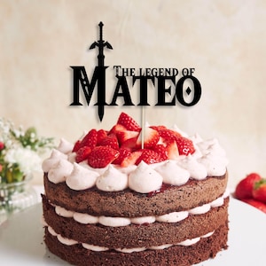 Puede incluir: Un adorno negro para pastel con el texto THE LEGEND OF MATEO y una espada integrada en la letra M. El adorno está colocado sobre un pastel de chocolate de tres capas decorado con glaseado blanco y fresas frescas en rodajas.