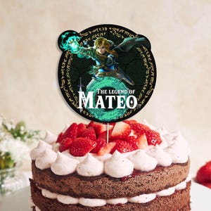 Puede incluir: Un adorno redondo para pastel con un personaje de "The Legend of Zelda" y el texto "The Legend of Mateo". El adorno está colocado sobre un pastel de chocolate decorado con crema de mantequilla blanca y fresas frescas.