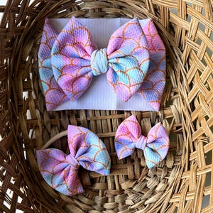 May include: Three mermaid-themed hair bows in a wicker basket. The largest bow is pink, blue, and gold with a scale pattern. The two smaller bows are the same pattern but in a smaller size.
