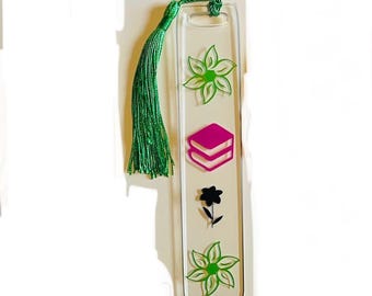 Peut inclure: Marque-page en acrylique transparent avec un pompon vert. Le marque-page présente des motifs floraux verts et une fleur noire, ainsi qu'un dessin de livre rose. Le pompon est fait de fil vert.