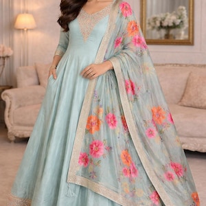 May include: A light blue Anarkali dress with a V-neckline, embellished with delicate beadwork. The dress features a matching floral print dupatta with pink and orange flowers. The hemline and dupatta border are adorned with intricate embroidery, creating an elegant ensemble.