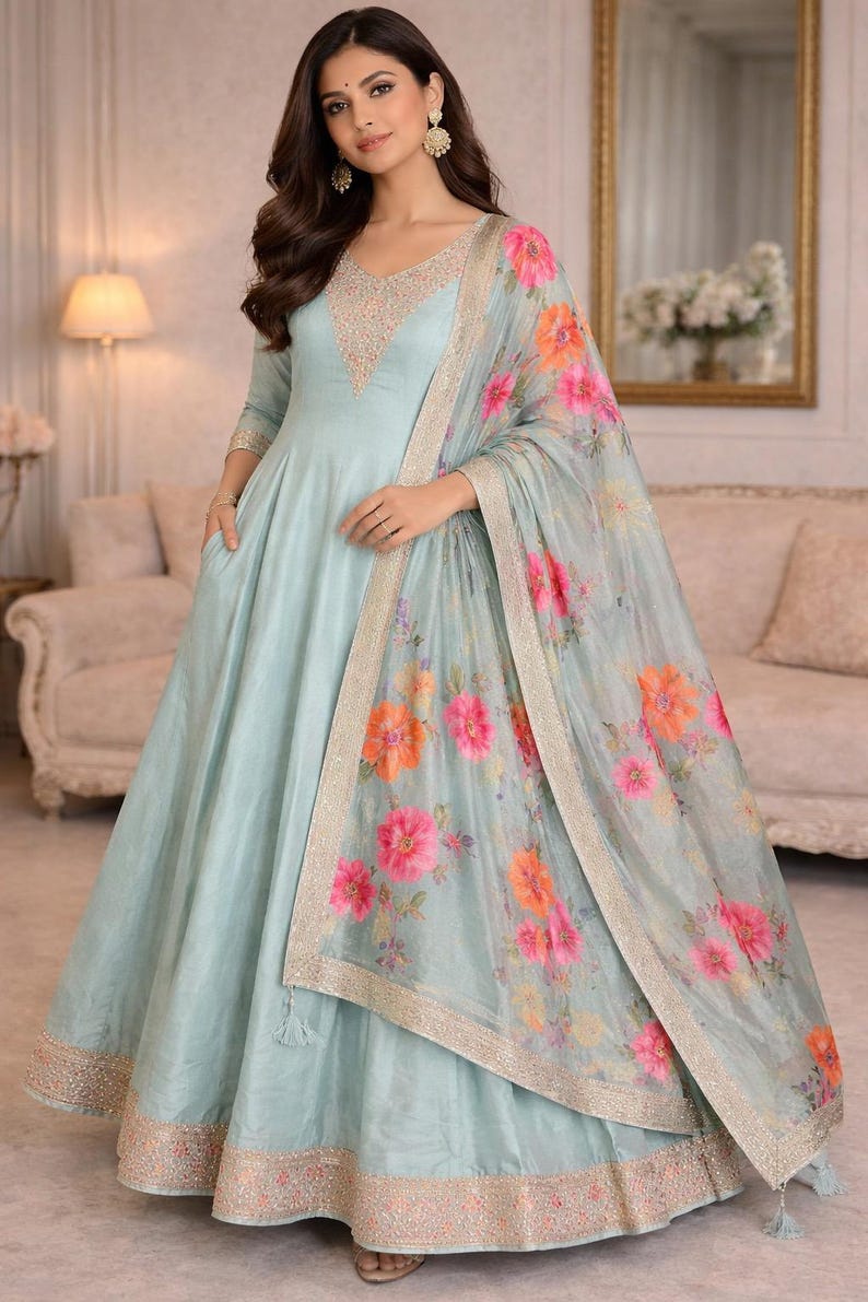 May include: A light blue Anarkali dress with a V-neckline, embellished with delicate beadwork. The dress features a matching floral print dupatta with pink and orange flowers. The hemline and dupatta border are adorned with intricate embroidery, creating an elegant ensemble.