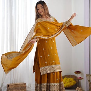 May include: Mustard yellow Indian salwar kameez outfit. The ensemble features a long tunic, wide-legged trousers, and a matching scarf. The tunic and trousers are decorated with detailed gold embroidery and small embellishments. The scarf has a decorative border.