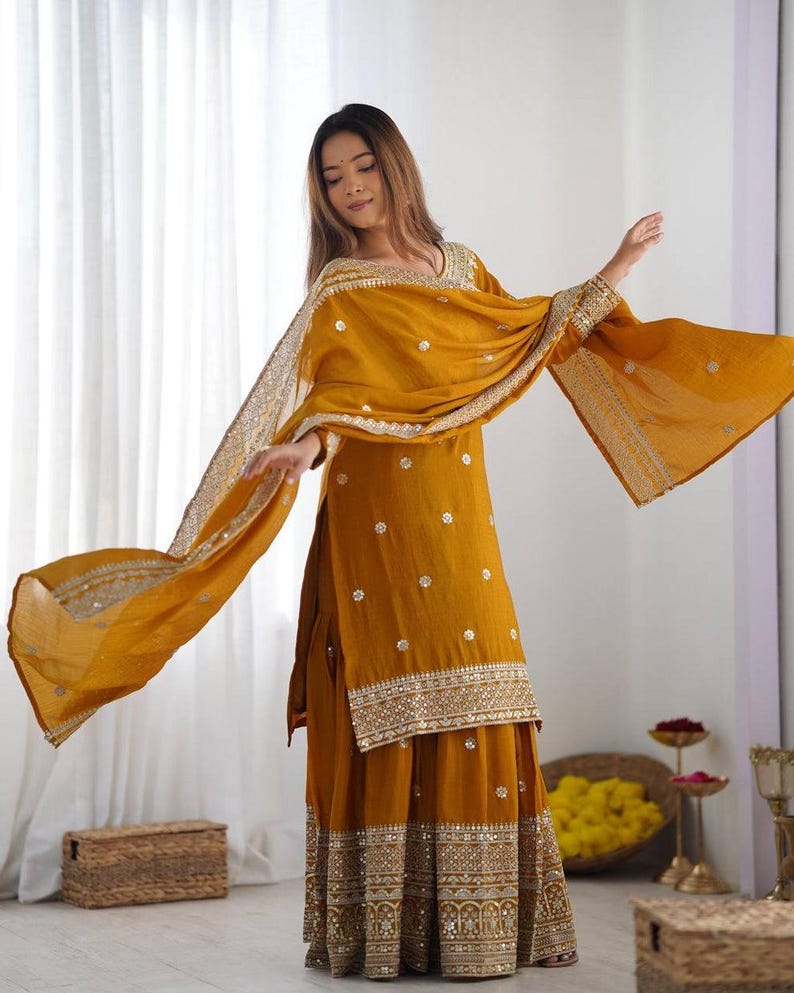 May include: Mustard yellow Indian salwar kameez outfit. The ensemble features a long tunic, wide-legged trousers, and a matching scarf. The tunic and trousers are decorated with detailed gold embroidery and small embellishments. The scarf has a decorative border.