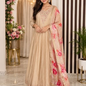 May include: A beige-colored, long-sleeved dress with intricate embroidery around the neckline and hem. A matching floral-print scarf with pink and red flowers drapes over the shoulder. The dress has a flowing, floor-length design, suitable for formal occasions.