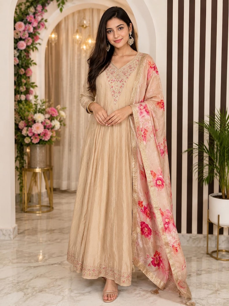 May include: A beige-colored, long-sleeved dress with intricate embroidery around the neckline and hem. A matching floral-print scarf with pink and red flowers drapes over the shoulder. The dress has a flowing, floor-length design, suitable for formal occasions.