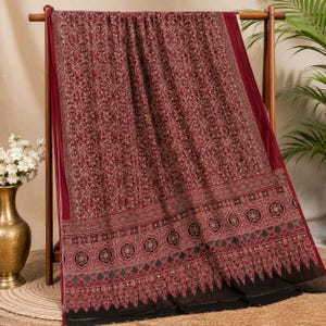 May include: A rectangular textile featuring a deep burgundy base with an intricate, repeating white and black geometric pattern across the main body. The lower section displays a wide, ornate border with circular motifs and pointed edges.
