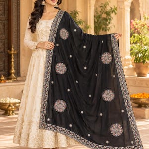 May include: A cream-colored floor-length dress with gold embroidery paired with a black shawl featuring circular multicolored floral patterns and a decorative border. The ensemble includes long sleeves and a flared skirt silhouette.