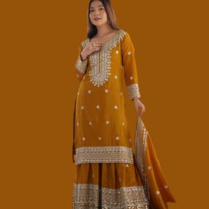 May include: A mustard-coloured salwar kameez outfit with silver embroidery. The long-sleeved tunic has detailed embellishments around the neckline and cuffs, paired with wide-legged trousers and a matching dupatta. The outfit is decorated with small, scattered embellishments.
